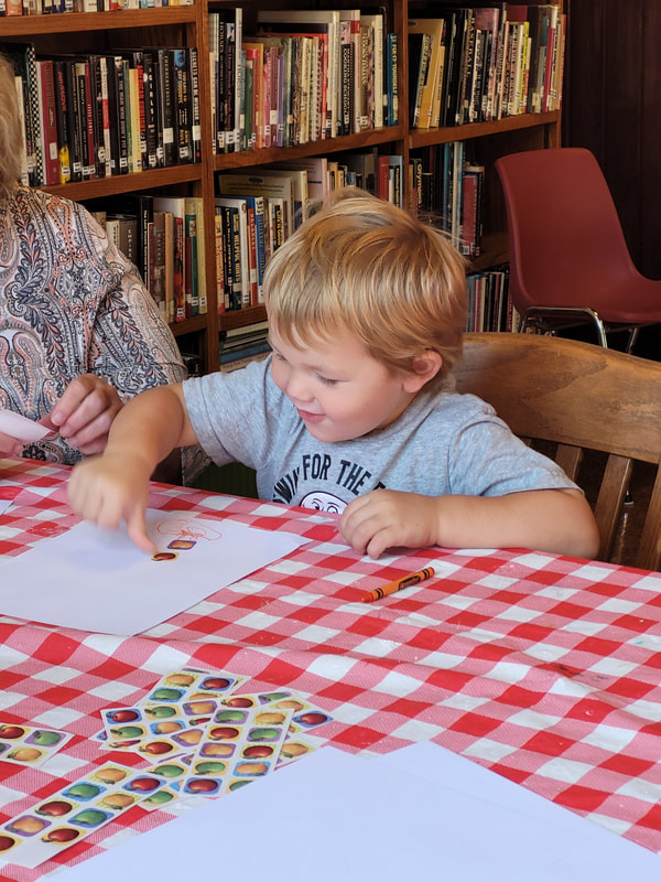 Young boy with blond hair sits at a table with red and white checked tablecloth. The boy is using his thumb to press a sticker onto a piece of paper.