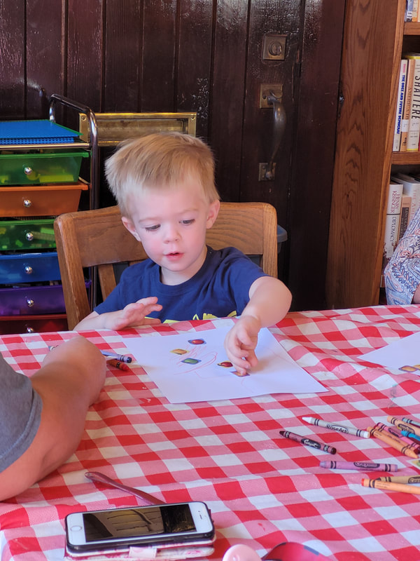 Young boy with blond hair and a blue shirt sits at a table with red and white checked tablecloth and presses stickers onto a piece of paper.