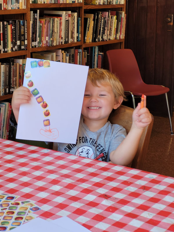 Smiling young boy with blond hair and a gray shirt sits at a table and holds up a paper with a line of stickers on it.