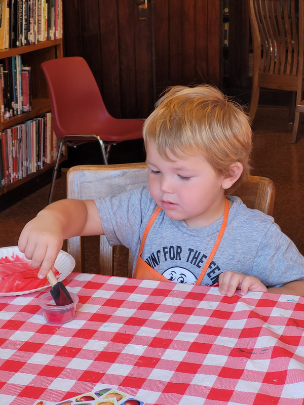 Young boy in a gray shirt and orange apron dips a sponge brush into a cup of red paint.