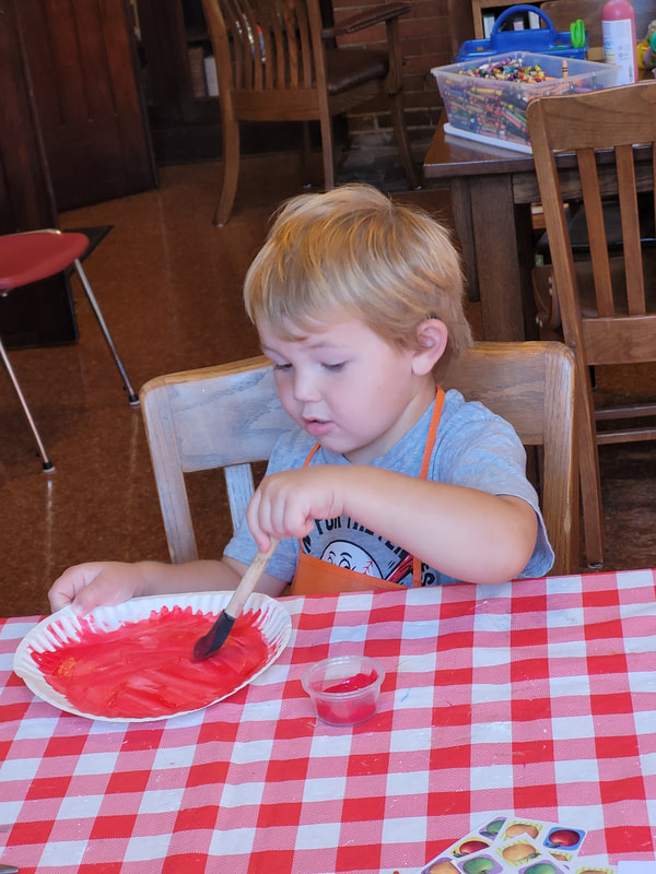 Young boy in a gray shirt and orange apron paints a paper plate with red.