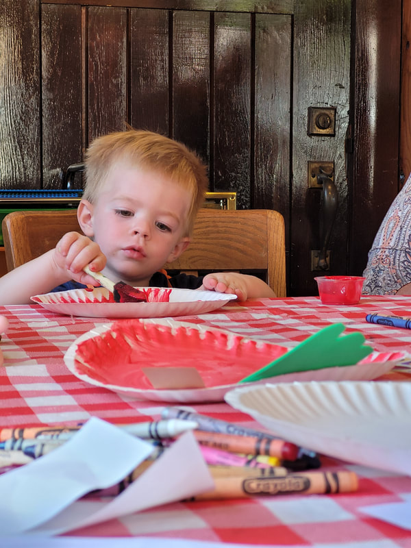 Young boy paints a paper plate red. Slightly blurry in the foreground, another red paper plate with green foam in the shape of a leaf sits on the table.